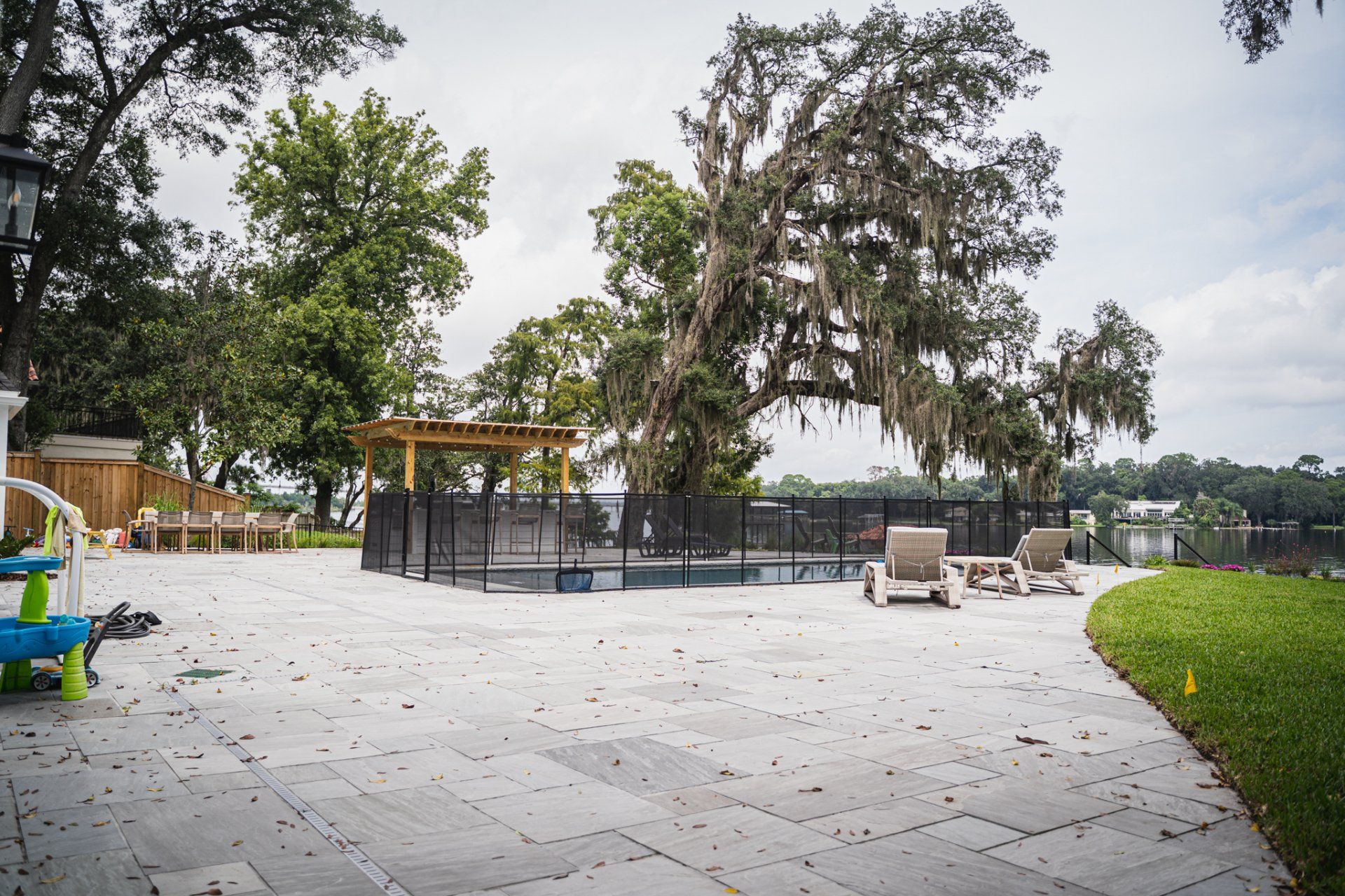 A pool area with patio, loungers, and a fenced pool, under trees near a body of water on a cloudy day.