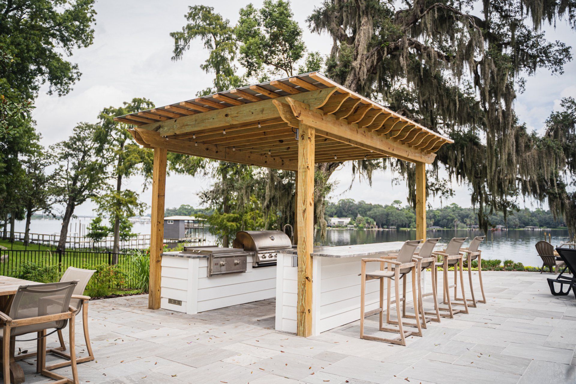 Outdoor kitchen with a grill, bar, and pergola overlooking a lake.