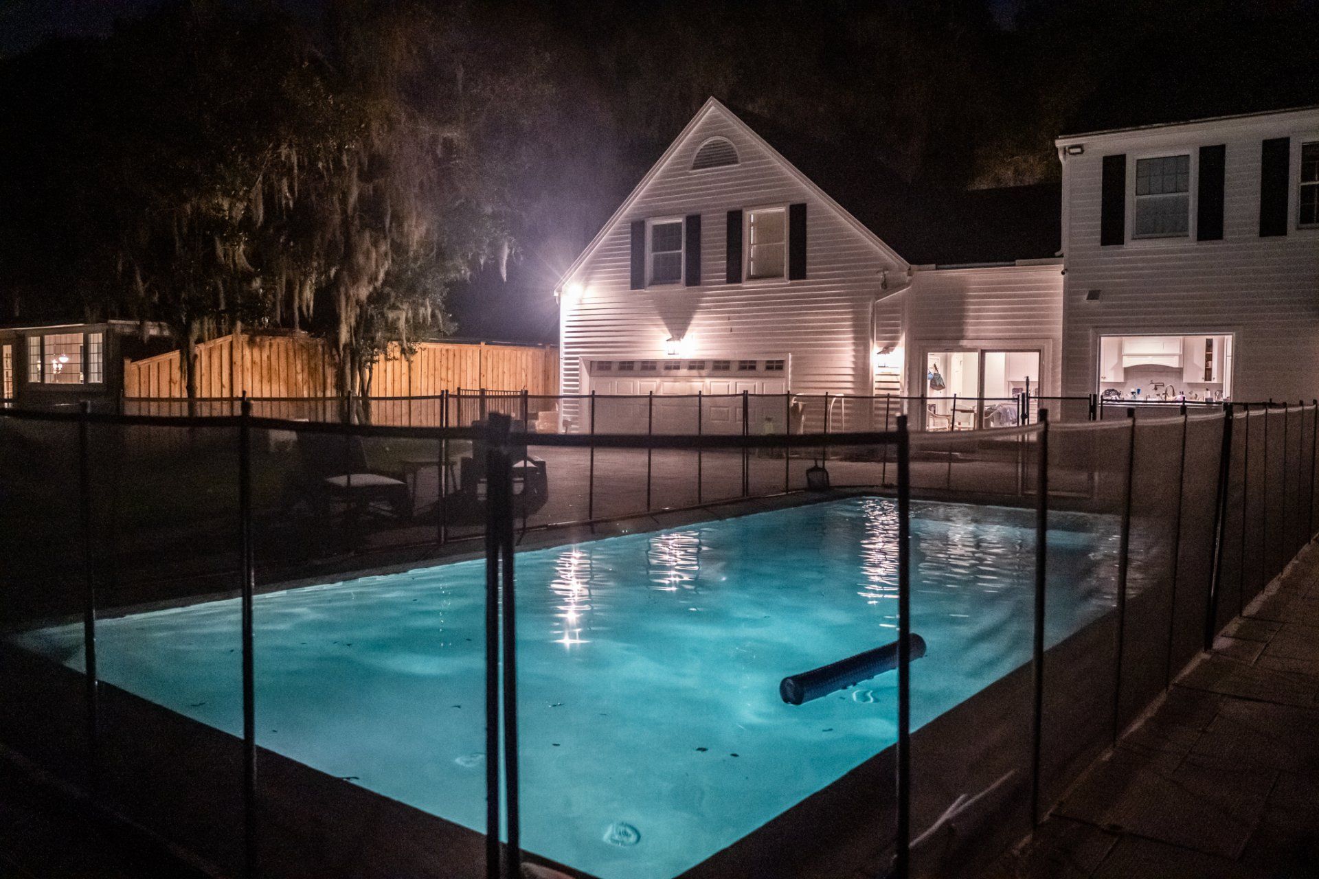 Night view of a lit swimming pool with a house in the background. A black fence surrounds the pool.