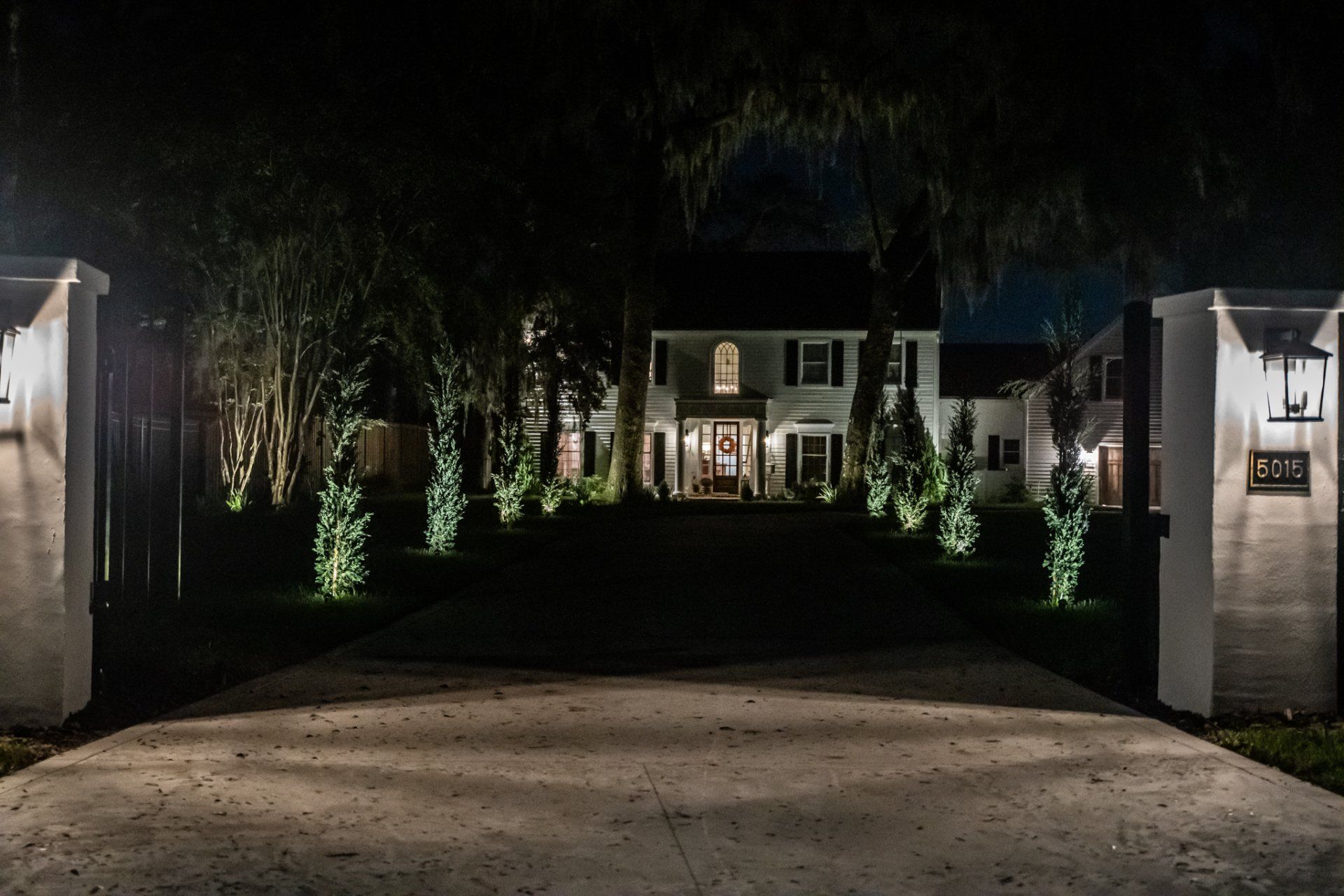 Night view of a house with a lit pathway, driveway, and pillars, with lights illuminating trees.