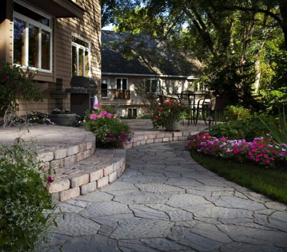 Stone path leads past steps, flower beds, and a patio area to a house with trees.