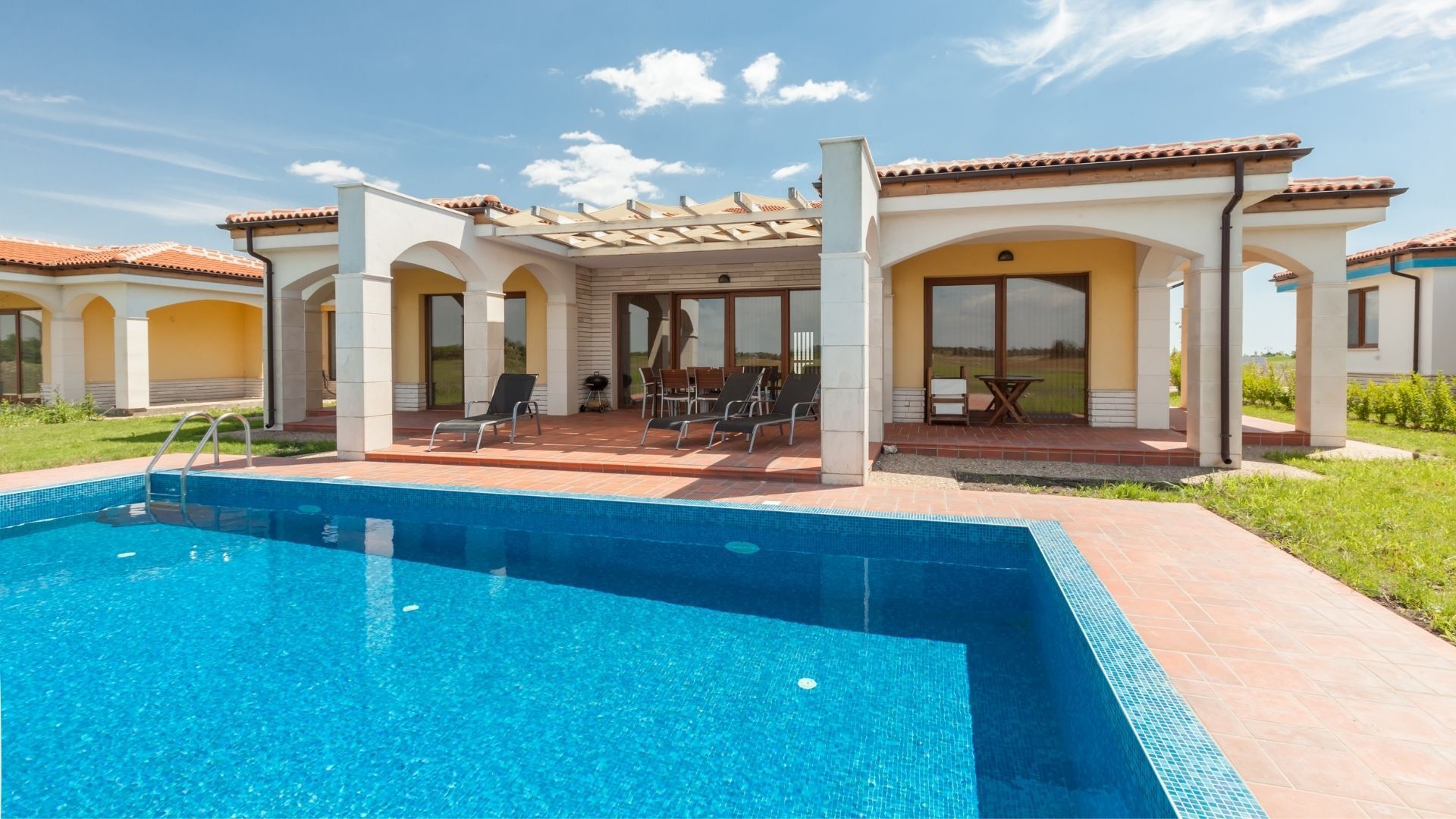 Pool with a villa in the background, blue water, red-tiled patio, sunny day.