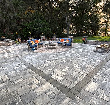 Patio with gray pavers, seating area with blue couches, fire pits, and trees in the background.