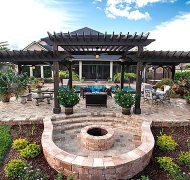 Outdoor patio with fire pit, pergola, and pool, brickwork, green plants, and blue sky.