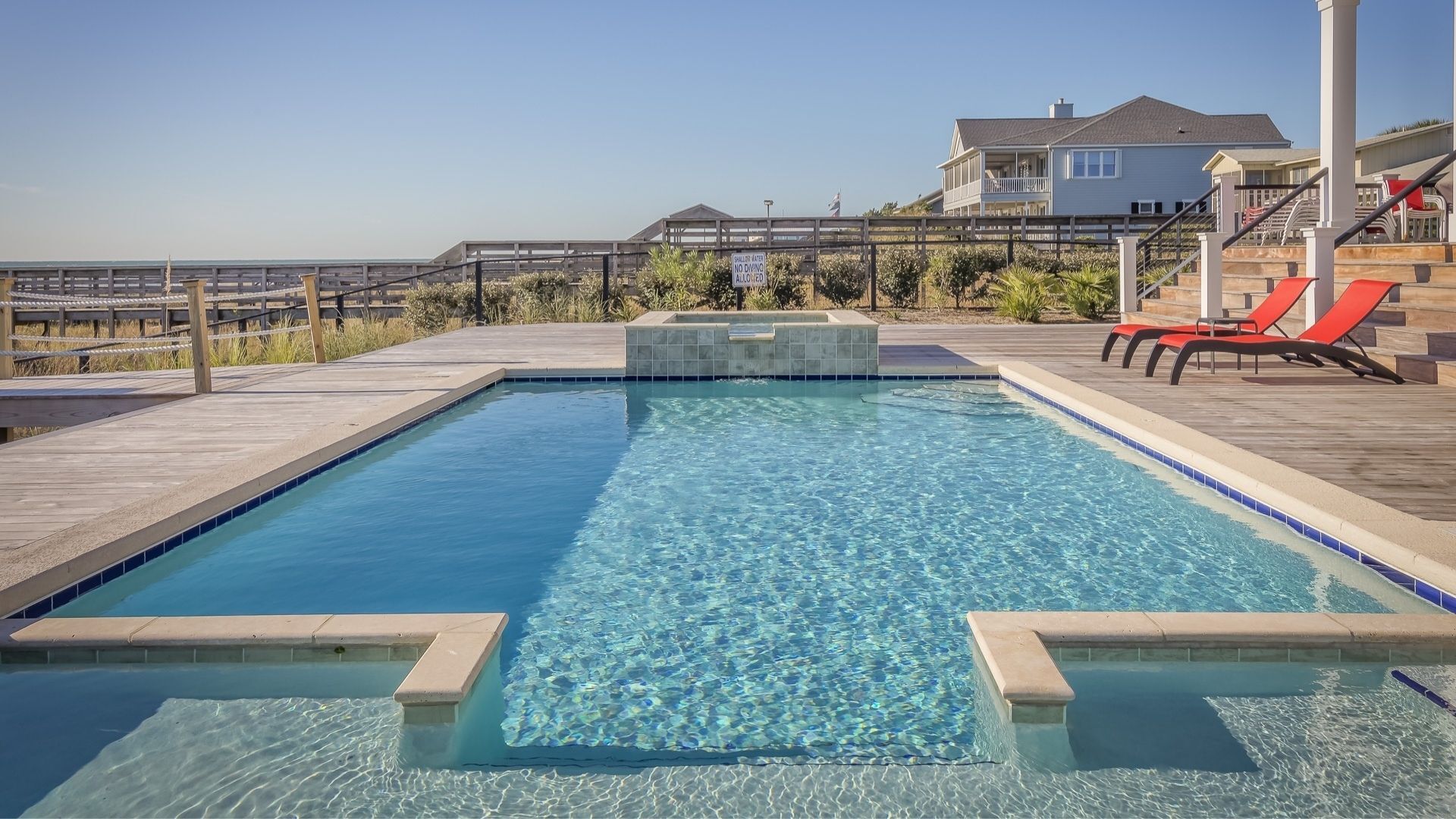 Rectangular swimming pool with light blue water and stone deck, overlooking the ocean. Two red lounge chairs sit by stairs.