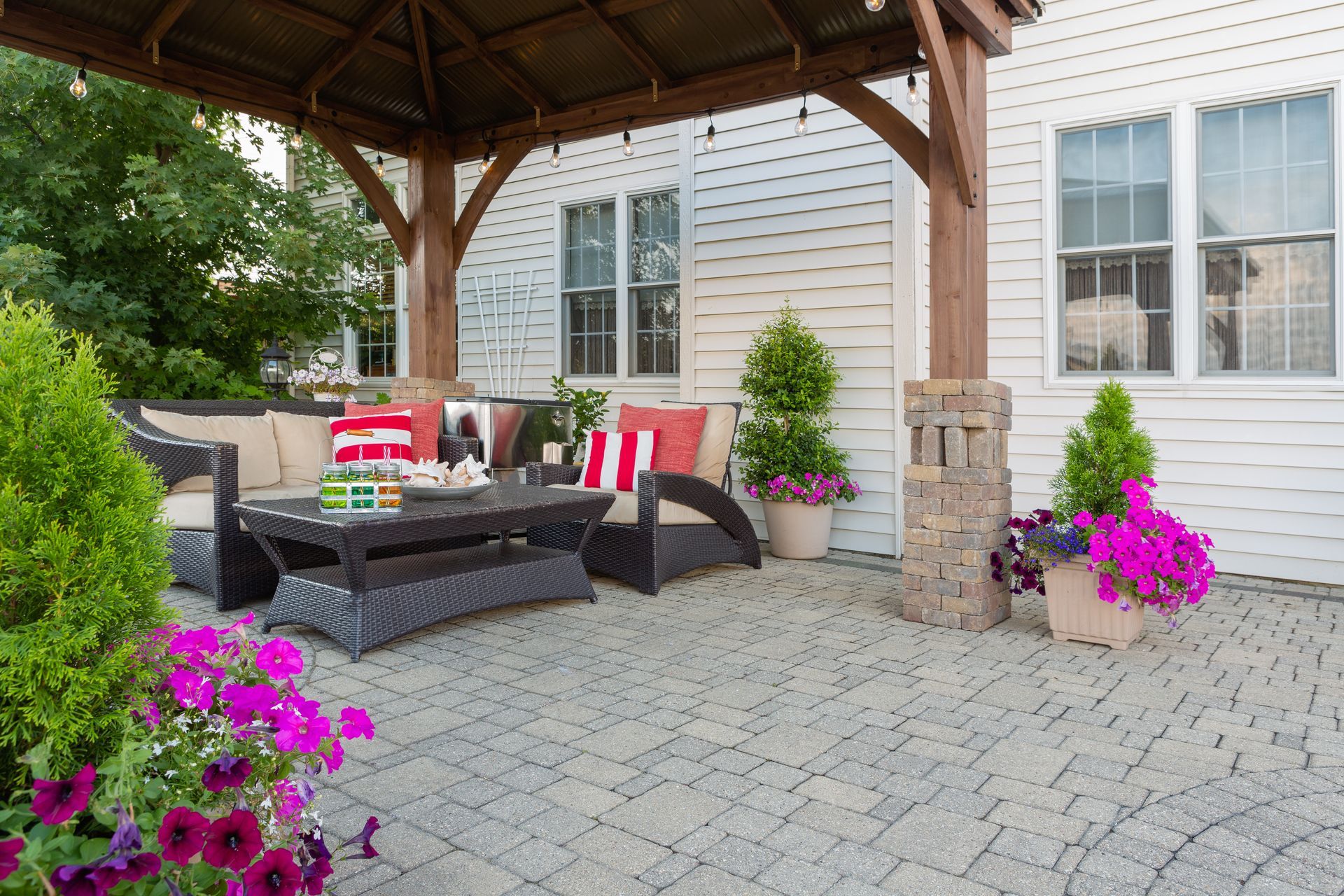 Patio with wicker furniture under a wooden gazebo, surrounded by flowers.