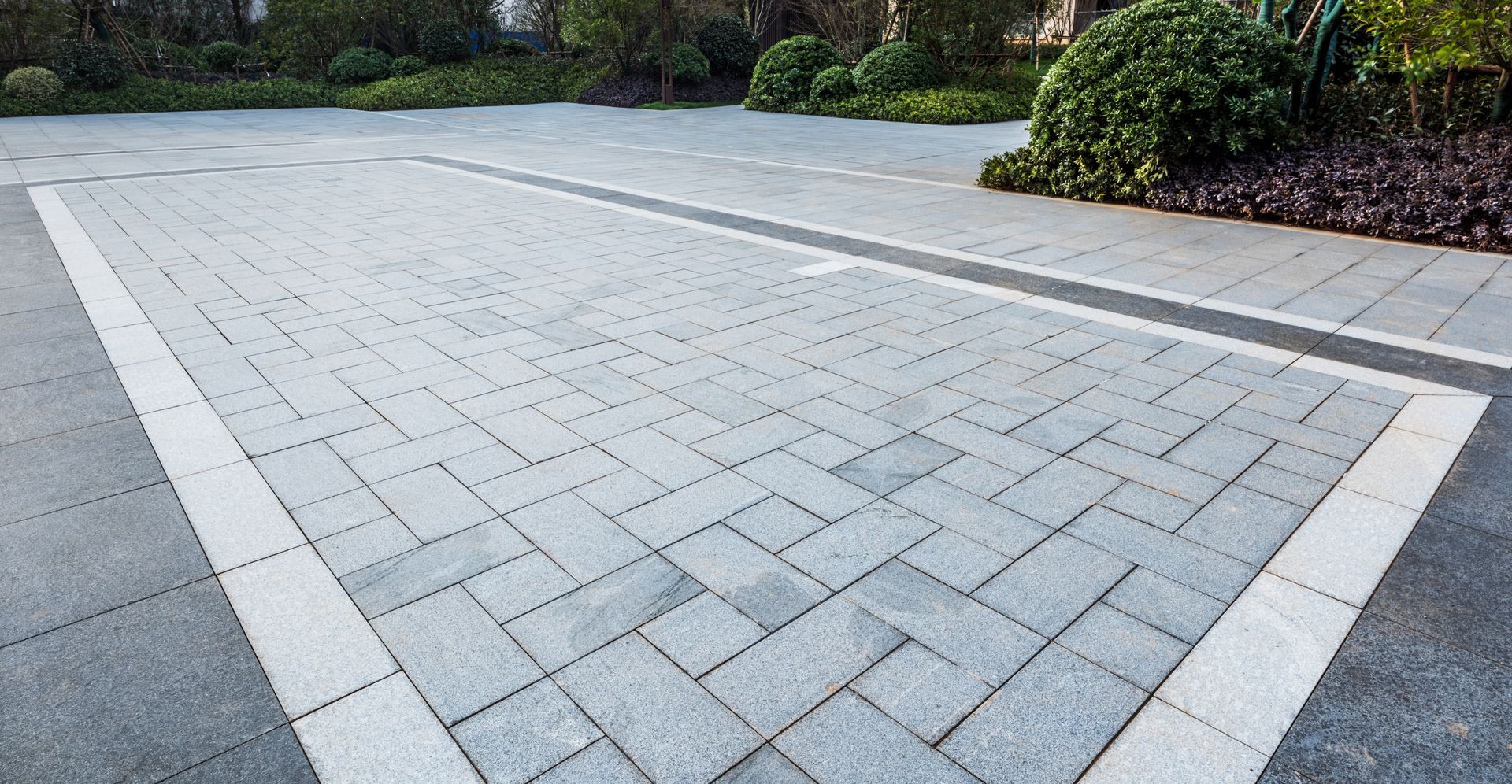 Gray brick paved area with light border and greenery in the background.