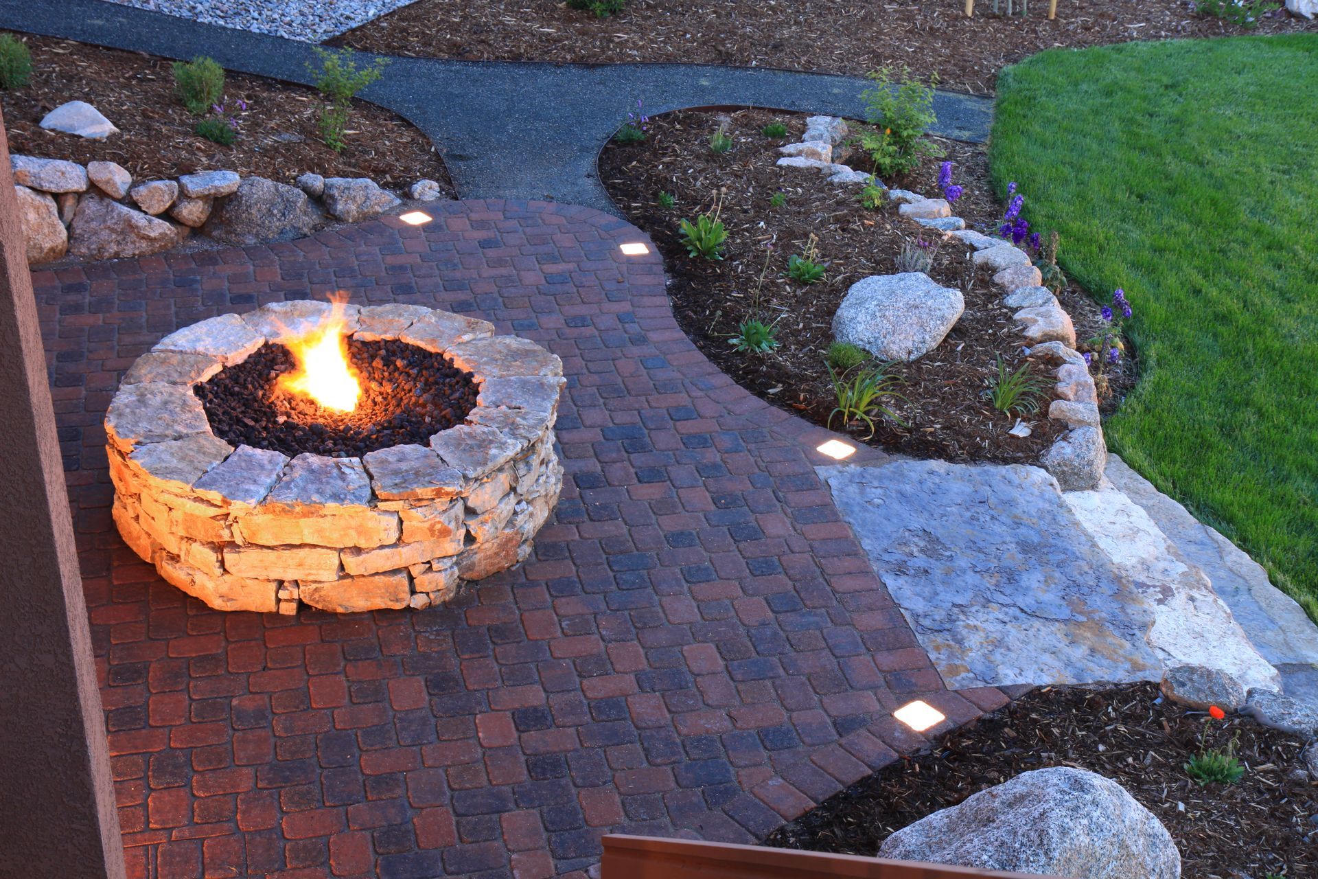 Stone fire pit ablaze on brick patio, surrounded by landscaped garden with built-in lights.