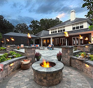 Stone patio with fire pit, torches, seating, and a house in the background at dusk.